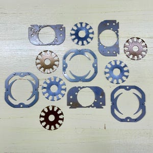 May include: A collection of various metal cutouts in shades of silver and bronze, scattered on a light wooden surface. The shapes include circles, squares, and starburst designs, with visible wear and tear.