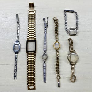 Może przedstawiać: Kolekcja zabytkowych elementów zegarków, w tym tarcze i paski. Paski są w odcieniach złota i srebra, z różnymi stylami ogniw. Niektóre mają puste prostokątne lub okrągłe tarcze zegarków.