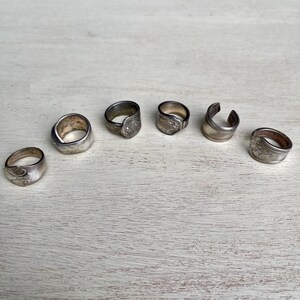 Puede incluir: Seis anillos de plata de diferentes diseños se colocan sobre una superficie de madera clara. Los anillos parecen estar hechos de cubiertos de plata, con detalles visibles como patrones florales y bandas curvas. Los anillos son de diferentes tamaños y estilos.