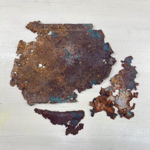Peut inclure: Quatre morceaux de métal rouillé de forme irrégulière avec une surface texturée. Le métal est principalement brun avec des taches de turquoise et de bleu-vert. Les bords sont dentelés et inégaux, avec des trous et des fissures.