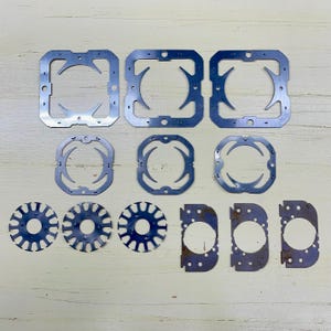Puede incluir: Un surtido de recortes metálicos plateados, que incluyen marcos cuadrados con diseños interiores curvos, formas circulares y piezas semicirculares. Los recortes están dispuestos sobre una superficie de madera de color claro.