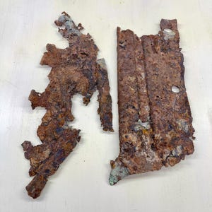 Peut inclure: Deux morceaux de métal fortement rouillé, aux formes irrégulières. Le métal est principalement dans les tons de brun et de rouille, avec quelques zones présentant des reflets verts et bleus. Les pièces semblent être des fragments d'un objet plus grand, peut-être d'un contexte industriel ou historique.