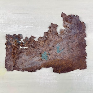 Peut inclure: Fragment de métal rouillé de forme irrégulière, aux teintes marron, violet et turquoise. La surface est rugueuse et texturée, avec des signes de dégradation et de petits trous, suggérant l'âge et l'exposition aux éléments.