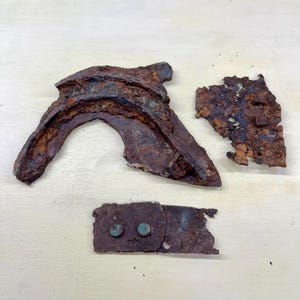 Peut inclure: Trois fragments de métal fortement corrodés et rouillés. Une pièce est incurvée, une autre est fragmentée et la troisième est rectangulaire avec deux rivets. Le métal présente une surface rugueuse et texturée avec des nuances de brun et de vert.