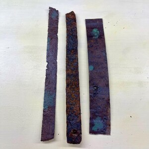 Peut inclure: Trois bandes de métal rectangulaires et patinées, de couleur brun rouille et violet. Des taches d'oxydation de couleur turquoise sont visibles. Les bandes varient en largeur et ont des bords irréguliers, suggérant l'âge et l'utilisation.