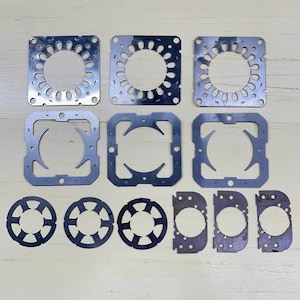 May include: A collection of various metal stamped parts, including square and circular shapes with intricate cut-out designs. The parts are arranged on a light-colored surface, showcasing their industrial aesthetic and precision.