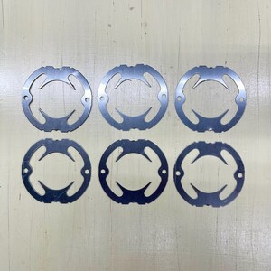 May include: Six silver-colored, circular metal pieces with a unique cut-out design. Each piece has a series of curved openings and small holes, arranged in a symmetrical pattern. The pieces are laid out on a light-colored surface.