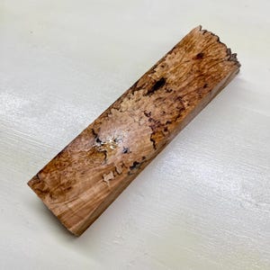 Könnte beinhalten: Ein rechteckiger Block aus poliertem Holz mit einem komplexen, wirbelnden Maserungsmuster in Schattierungen von hellem Beige, Goldbraun und Dunkelschokoladenbraun. Die Oberfläche zeigt natürliche organische Texturen, dunkle Adern und Astlöcher.