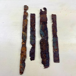 May include: Five weathered, rectangular metal pieces with a heavily rusted, textured surface. The pieces vary in length and width, displaying shades of brown, orange, and hints of blue-green. The edges are irregular, suggesting age and decay.