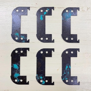 May include: Six black metal corner brackets with a distressed finish and turquoise accents. Each bracket has a right-angle shape with a curved corner, a small tab, and three pre-drilled holes. The brackets are arranged on a light-colored wooden surface.
