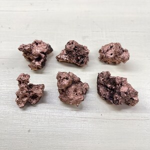Könnte beinhalten: Sechs Stücke rohes Kupfererz, die eine Vielzahl von Formen und Texturen aufweisen. Das Kupfer hat eine rotbraune Farbe mit dunkleren Bereichen, und die Stücke haben ein raues, poröses Aussehen. Die Objekte sind auf einer hellen Holzoberfläche angeordnet.