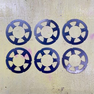Puede incluir: Seis arandelas metálicas circulares de color azul oscuro con un diseño recortado único. Cada arandela tiene un agujero central rodeado por un anillo con formas geométricas espaciadas uniformemente. Las arandelas están dispuestas sobre una superficie texturizada de color amarillo claro.