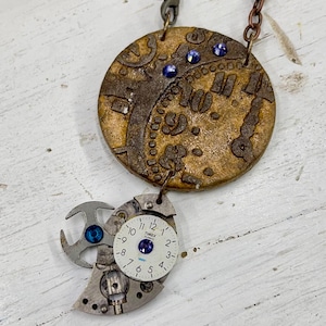 May include: A steampunk-style necklace featuring a circular pendant with a clock face design in shades of brown and gold, adorned with blue gems. Below, a cluster of watch parts and a Timex watch face add to the unique, vintage aesthetic.