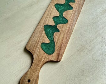 May include: A wooden serving board with a handle and a flowing river of green epoxy resin. The board is rectangular with a rounded handle and a hole for hanging. The wood has a natural grain, and the resin adds a decorative touch.