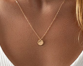 May include: A gold-toned necklace featuring a delicate chain and a small, circular pendant with a textured, hammered surface. The pendant hangs centrally from the chain, which rests against the collarbone area, providing a simple and refined look.