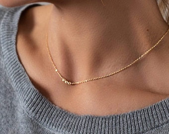 May include: A delicate gold chain necklace featuring three small, round, matte-finish gold beads positioned slightly off-center. The necklace rests against the neck, showcasing a fine link structure and a consistent metallic gold tone throughout the piece.