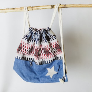 Op de afbeelding: Een rugzak met trekkoord, met een denimblauwe onderkant en een patroon aan de bovenkant in rood, zwart en wit. De tas heeft een witte sterapplicatie en witte touwriemen. Hij hangt aan een natuurlijke houten tak.