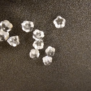 Könnte beinhalten: Eine Ansammlung klarer, blütenförmiger Ohrringverschlüsse. Die Ohrringverschlüsse bestehen aus einem durchscheinenden Material und liegen auf einer dunklen, strukturierten Oberfläche. Die Ohrringverschlüsse sind klein und filigran.