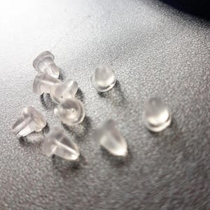 Op de afbeelding: Een close-up van een groep doorzichtige plastic oorbelruggen. De ruggen zijn klein en rond, met een lichte inkeping in het midden.