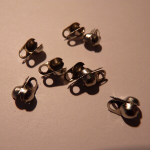 May include: A collection of small, silver-colored metal cord ends, scattered on a light-colored surface. Each cord end has a rounded, cup-like shape with a small loop for attaching to a cord or string. The metal has a shiny finish.