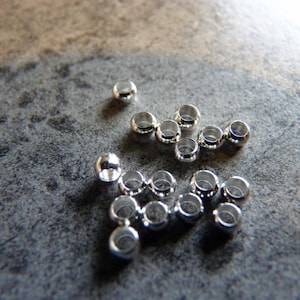 May include: A group of silver metal beads, some are round and some are slightly oval shaped.