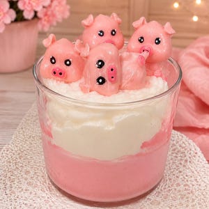 May include: A clear glass container filled with a layered pink and white substance, topped with four small, glossy pink pig figurines. The pigs have black eyes and pink snouts, appearing to rest on a textured white layer resembling whipped cream.