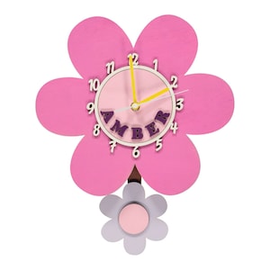 May include: A pink flower-shaped wall clock with a light pink clock face and yellow hands. The clock has the name "AMBER" in purple letters. A smaller grey and pink flower hangs below.