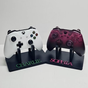 May include: Two custom-designed video game controller stands with personalised names "Charlie" and "Sophia". The stands are black and the controllers are white and purple.