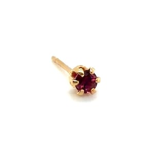 May include: A single gold stud earring with a round, deep red gemstone. The gemstone is set in a six-prong setting. The earring post is thin and gold. The earring is set against a white background.
