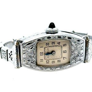 May include: A silver-toned Bulova wristwatch featuring an ornate, engraved metal case and matching link bracelet. The rectangular watch face has a cream-colored background with black Arabic numerals and dark blue hands, topped with a black crown.