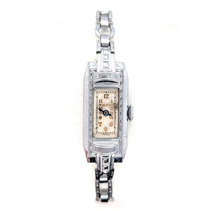 May include: A silver-toned Bulova wristwatch featuring a rectangular face with a cream-colored dial, black Arabic numerals, and black hands. The watch case has an intricate engraved border and is attached to a matching silver-toned metal link bracelet.