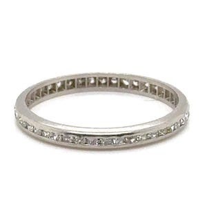 May include: A thin silver band with a row of small, square-shaped diamonds set in a continuous line around the entire band.
