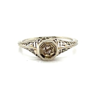 May include: A vintage ring with an octagonal central stone and detailed filigree work. The ring has a small, round, pale gemstone set in a white metal band. The design is symmetrical and delicate, with an antique aesthetic.