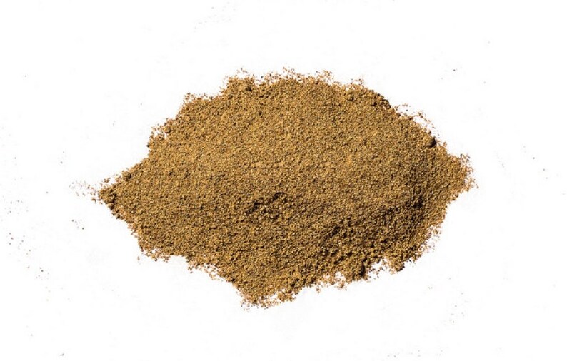 May include: A mound of finely ground, light brown powder on a white backdrop. The powder has a slightly coarse texture, suggesting it is a spice or herb. The image is well-lit, showing the details of the powder.