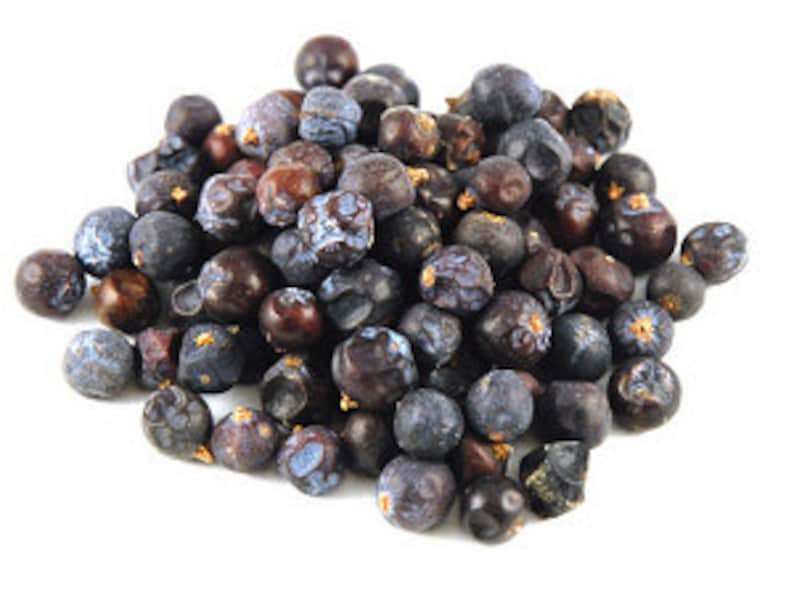 May include: A close-up of a pile of dried juniper berries. The berries are a mix of dark blue and brown, with a wrinkled texture. They are clustered together against a white backdrop. Juniper berries are a key ingredient in gin and are used in cooking.