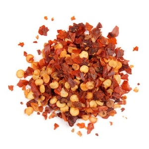 May include: A close-up of a pile of red pepper flakes and seeds. The flakes are dried and have a slightly orange hue. The seeds are small and round.