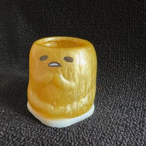 May include: A yellow ceramic cup with a white rim and a cartoon face with black eyes and a mouth. The cup has a shiny, metallic finish.
