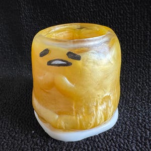 May include: A gold-colored ceramic pencil holder shaped like a cartoon character with a white base. The character has a black outline and a simple face with a frown.