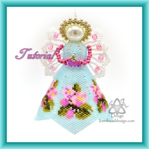 以下が含まれることがあります： 薄い青色のドレスを着たビーズ製の天使のオーナメント。ピンクの花とピンクのビーズのハローが付いています。天使はピンクのビーズのネックレスを手に持っています。オーナメントには「チュートリアル」と「Iceni Bead Design」のラベルが付いています。
