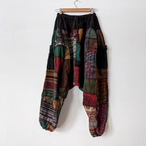 Może przedstawiać: Spodnie w stylu patchwork z czarną talią i sznurkiem. Spodnie posiadają r&oacute;żnorodne kolorowe łaty materiału w odcieniach czerwieni, zieleni, brązu i fioletu. Spodnie wiszą na drewnianym wieszaku na białej ścianie.