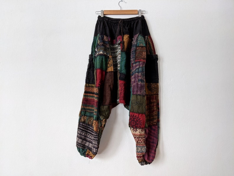 Może przedstawiać: Spodnie w stylu patchwork z czarną talią i sznurkiem. Spodnie posiadają r&oacute;żnorodne kolorowe łaty materiału w odcieniach czerwieni, zieleni, brązu i fioletu. Spodnie wiszą na drewnianym wieszaku na białej ścianie.