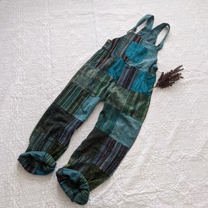 May include: Blue and green patchwork overalls with adjustable straps and pockets. The overalls feature a variety of patterned fabric squares in shades of teal, navy, and forest green. The cuffs are rolled up, and a sprig of dried lavender rests nearby.