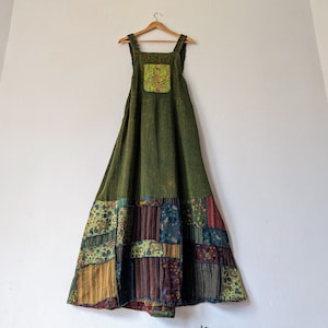 Pode incluir: Vestido longo verde-oliva com detalhes de patchwork na bainha e um bolso floral. O vestido tem alças ajustáveis e está pendurado em um cabide de madeira. O patchwork inclui vários tecidos florais e estampados.