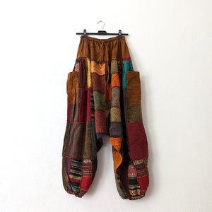 Może przedstawiać: Wielokolorowe spodnie w stylu patchwork w odcieniach brązu, czerwieni, pomarańczy i zieleni wiszą na czarnym wieszaku. Spodnie mają luźny kr&oacute;j z wieloma łatami z tkaniny i dwiema dużymi kieszeniami. W pasie sznurek.