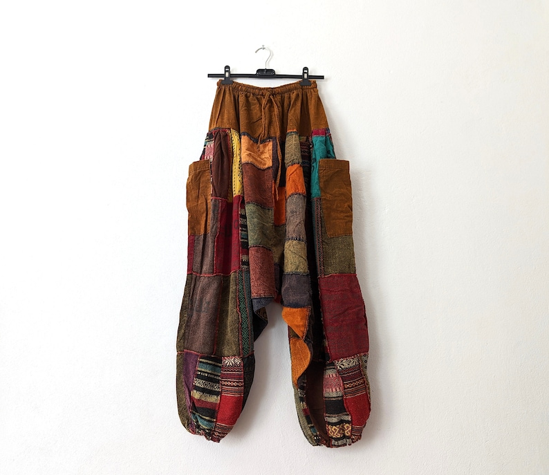 Może przedstawiać: Wielokolorowe spodnie w stylu patchwork w odcieniach brązu, czerwieni, pomarańczy i zieleni wiszą na czarnym wieszaku. Spodnie mają luźny kr&oacute;j z wieloma łatami z tkaniny i dwiema dużymi kieszeniami. W pasie sznurek.