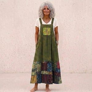May include: An olive green overall dress with a patchwork hem. The dress has a square bib pocket with a floral design and two front pockets. A white t-shirt is worn underneath. The model is barefoot.