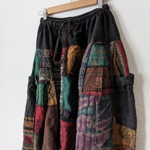Może przedstawiać: Spodnie w stylu patchwork z czarną talią i sznurkiem. Spodnie posiadają r&oacute;żnorodne kolorowe łaty materiału w odcieniach czerwieni, zieleni, brązu i fioletu. Spodnie wiszą na drewnianym wieszaku na tle białej ściany.