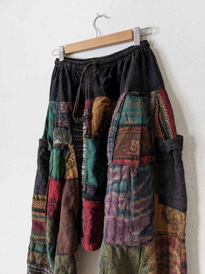 Może przedstawiać: Spodnie w stylu patchwork z czarną talią i sznurkiem. Spodnie posiadają r&oacute;żnorodne kolorowe łaty materiału w odcieniach czerwieni, zieleni, brązu i fioletu. Spodnie wiszą na drewnianym wieszaku na tle białej ściany.