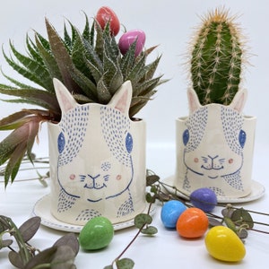 Puede incluir: Dos macetas de cerámica con forma de conejito, cada una con un platillo a juego. Una maceta contiene una suculenta, la otra un cactus. Los conejitos son blancos con detalles azules y rosas. Caramelos de huevo de Pascua de colores están esparcidos.