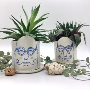 May include: Two white ceramic plant pots with a quirky face design in blue and pink, each containing a succulent plant. The pots sit on matching saucers, with a neutral background and decorative greenery.